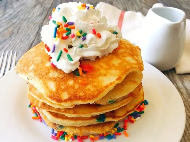 A stack of sweet cream pancakes topped with whipped cream and rainbow sprinkles.