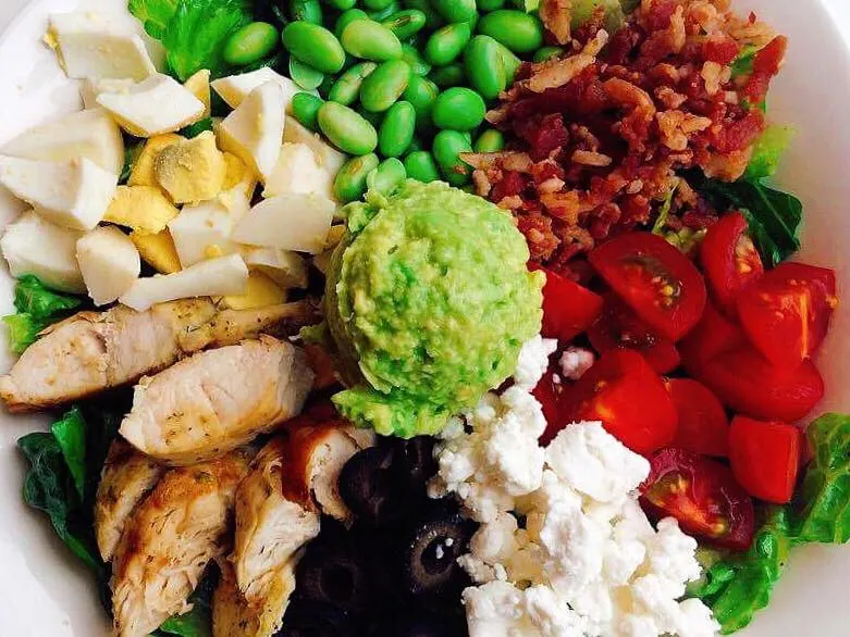 A white bowl with edamame, lettuce, bacon, tomatoes, feta cheese, avocado, black olives, chicken, and hard boiled eggs to make a Cobb salad.