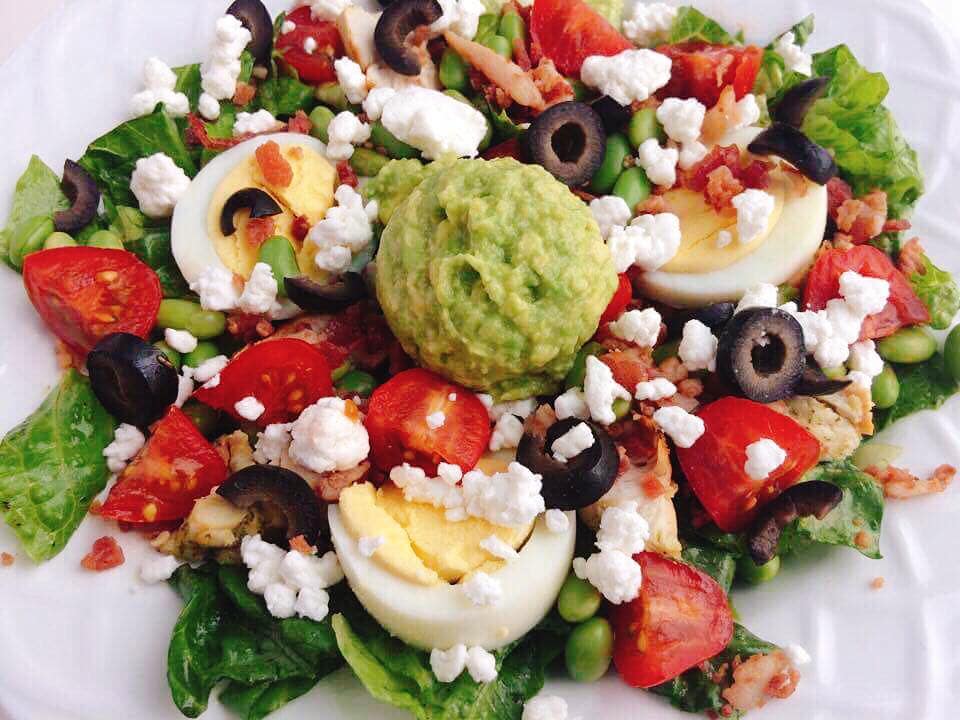 A white bowl filled with lettuce, tomatoes, olives, hard boiled eggs, avocado, bacon, and chicken to make a Cobb salad.