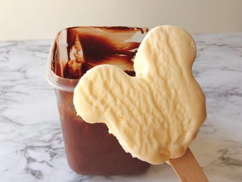 A vanilla Mickey Mouse shaped ice cream bar in front of a container of melted to chocolate to make Homemade Hand Dipped Ice Cream Bars