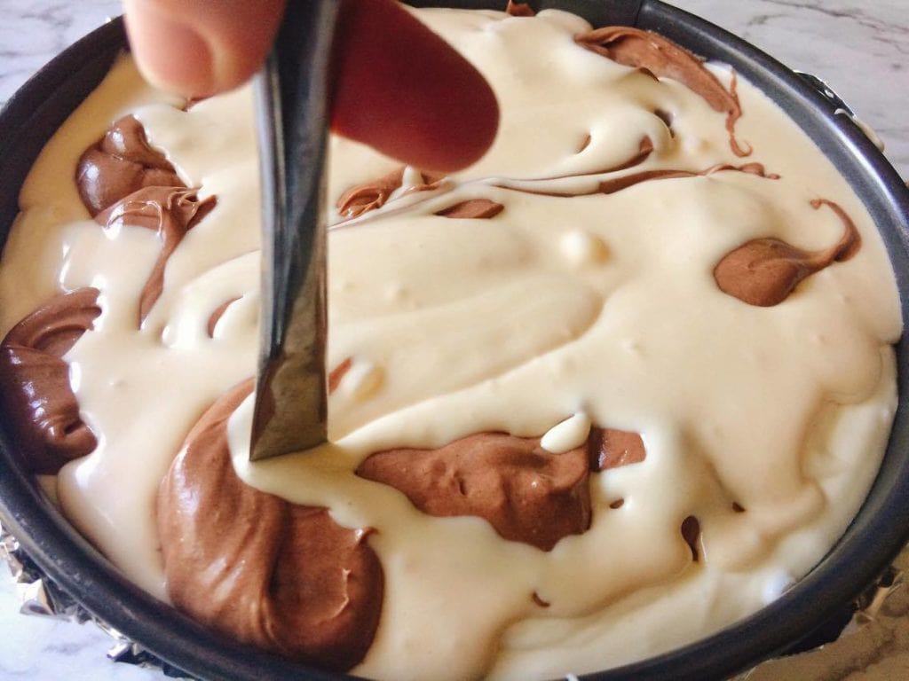 A knife swirling caramel, chocolate and classic cheesecake to make Caramel Pecan Turtle Cheesecake Copycat Recipe from Cheesecake Factory
