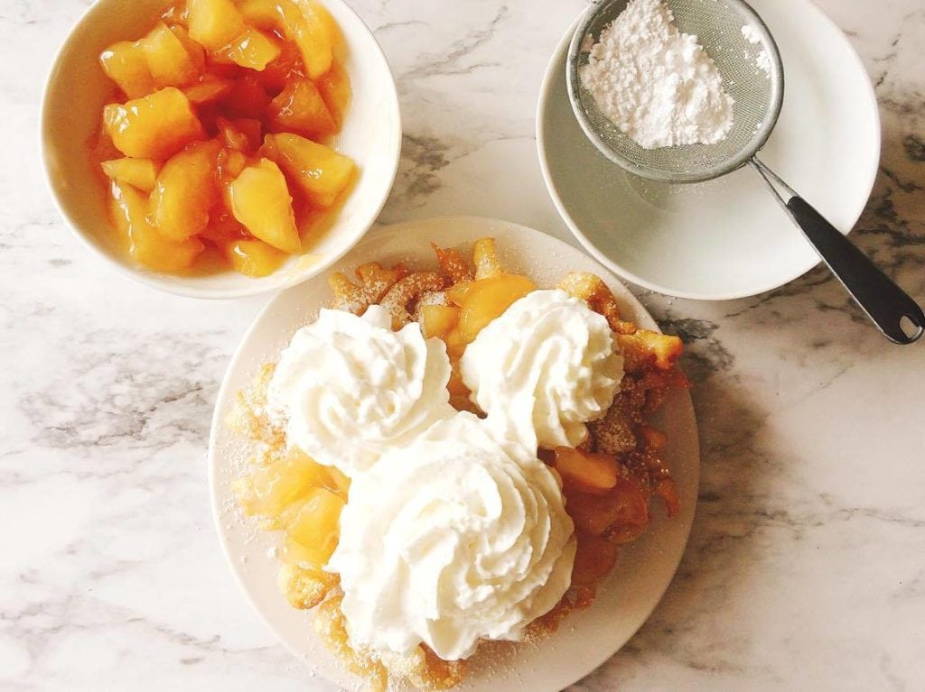 Disneyland's Apple Pie Funnel Cake on a plate with a whipped cream mickey head, a bowl of apple pie filling, and a bowl of powdered sugar.