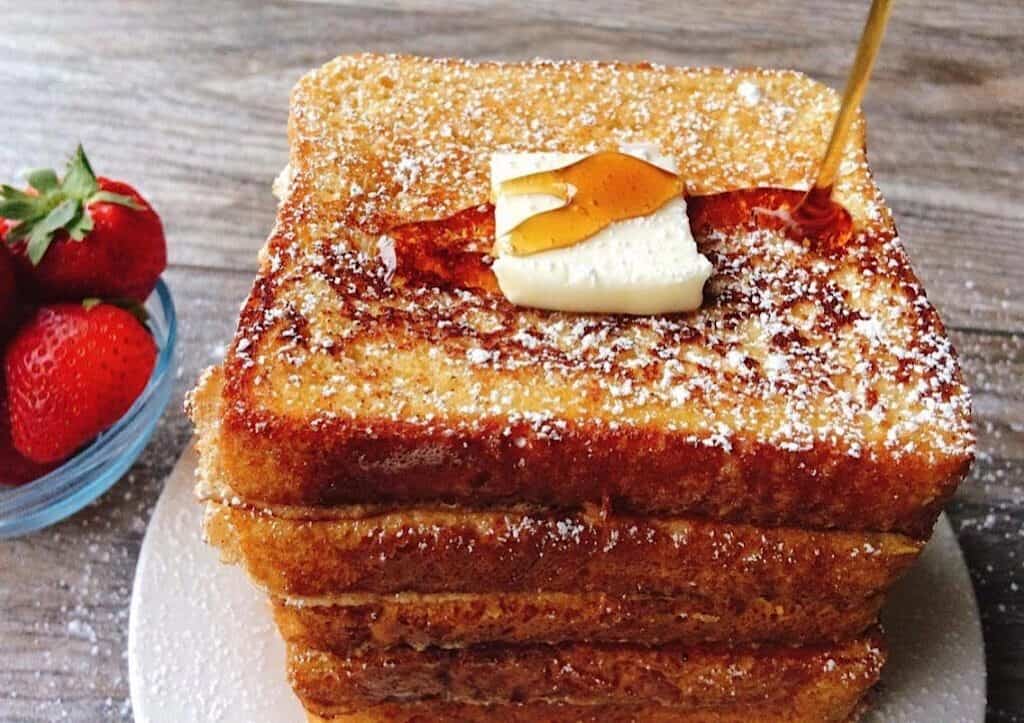 Syrup being poured on a stack of French Toast topped with a pat of butter and a side of strawberries.