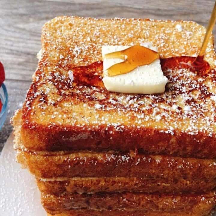 Syrup being poured on a stack of French Toast topped with a pat of butter and a side of strawberries.
