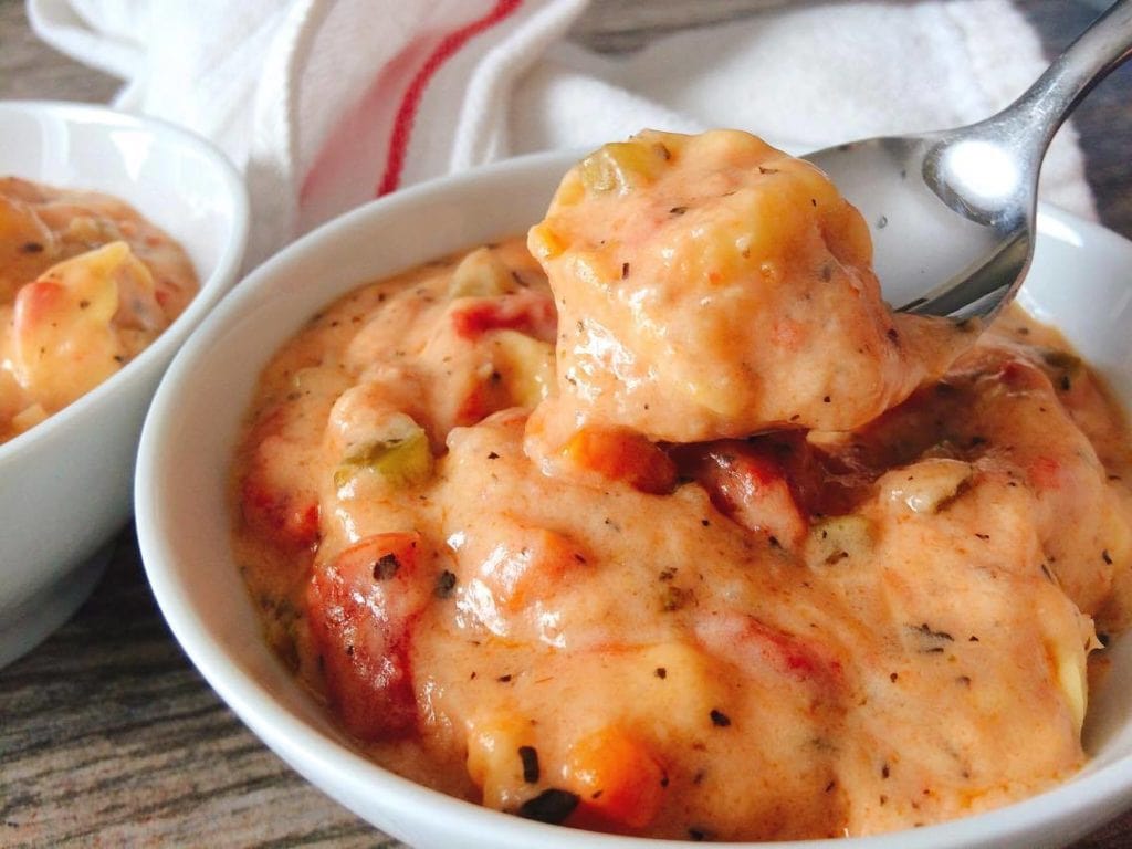 A spoon of Instant Pot Cheesy Tortellini Tomato Soup over. white bowl of soup with a kitchen towel in the background