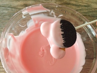 An Oreo on a stick with pink Minnie Mouse Ears over a bowl of melted pink chocolate.