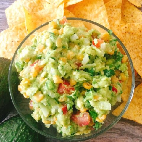 A bowl of Fully-Loaded Chunky Guacamole surrounded by tortilla chips and an avocado.