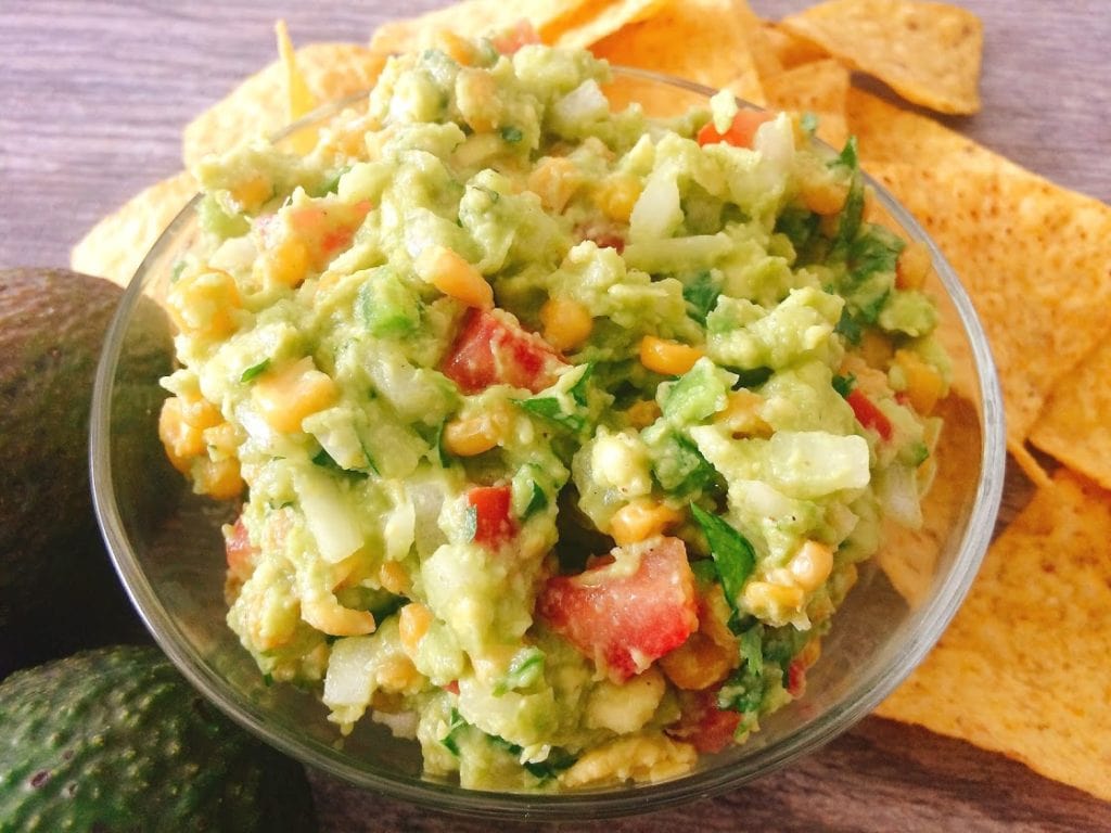 A bowl of Fully-Loaded Chunky Guacamole surrounded by tortilla chips and an avocado.