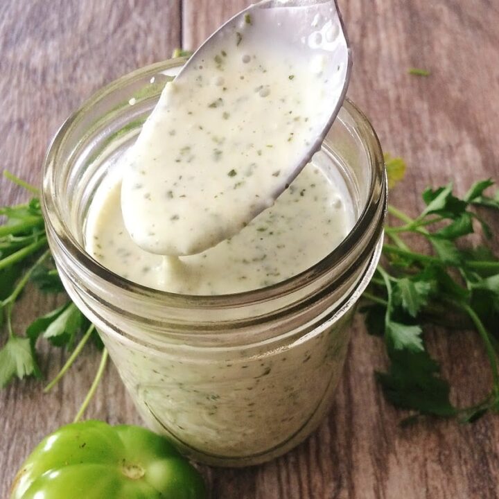 A spoon in a jar of creamy tomatillo ranch dressing surrounded by cilantro and a tomatillo.