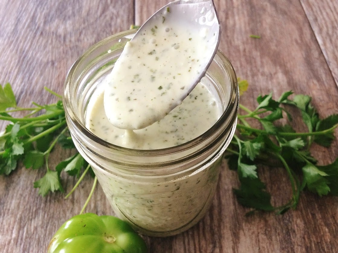 A spoon in a jar of creamy tomatillo ranch dressing surrounded by cilantro and a tomatillo.