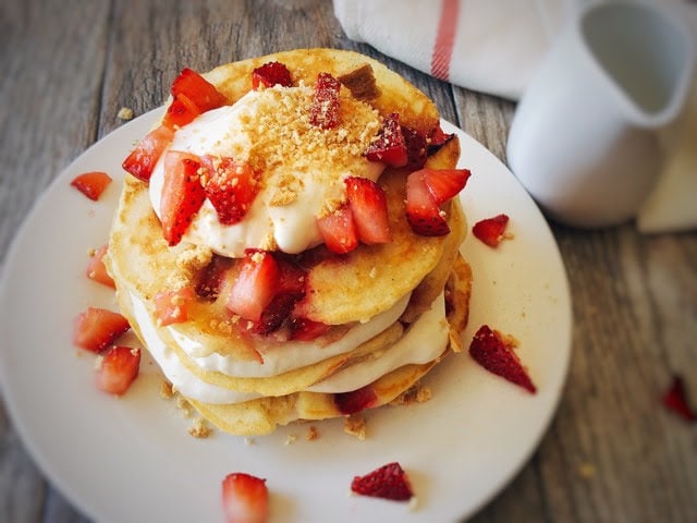 A stack of Strawberry Cheesecake Pancakes sprinkled with chopped strawberries on a white plate with a syrup cup and a kitchen towel.