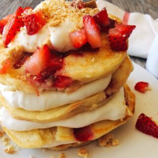 A stack Strawberry Cheesecake Pancakes sprinkled with chopped strawberries and layered with Cheesecake Topping on a white plate with a syrup cup and a kitchen towel in the background.