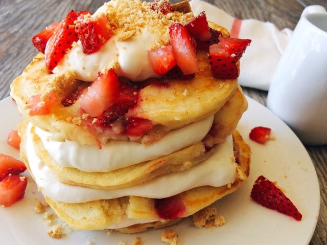 A stack Strawberry Cheesecake Pancakes sprinkled with chopped strawberries and layered with Cheesecake Topping on a white plate with a syrup cup and a kitchen towel in the background.