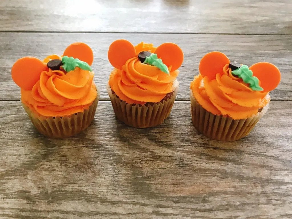 Three cupcakes decorated with orange frosting and orange candy melts to look like a Mickey Mouse Pumpkin to make Halloween Cupcakes.