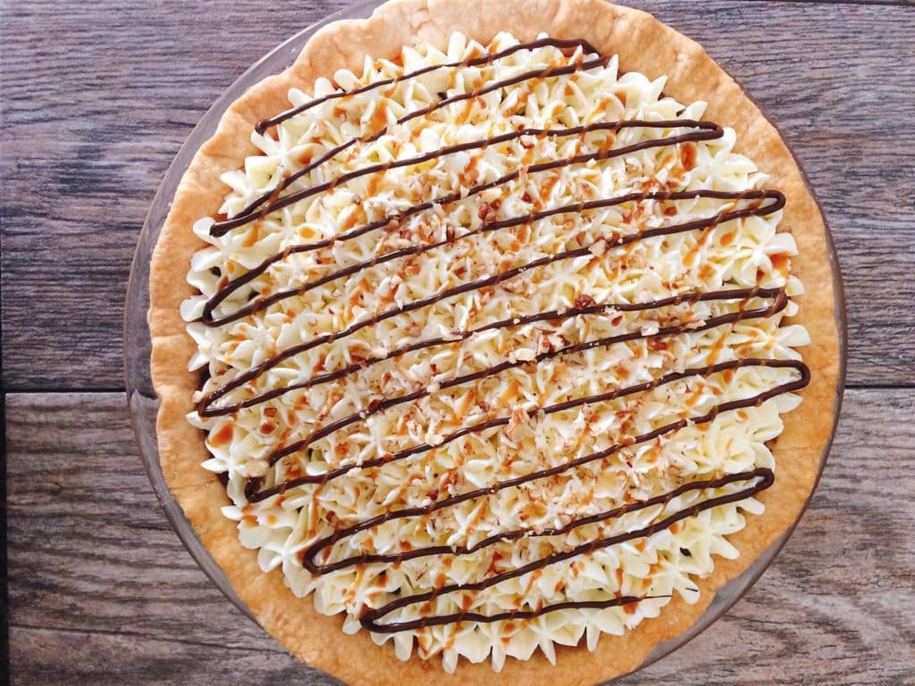 A Caramel Pecan Silk Pie drizzled with melted chocolate on a grey background.