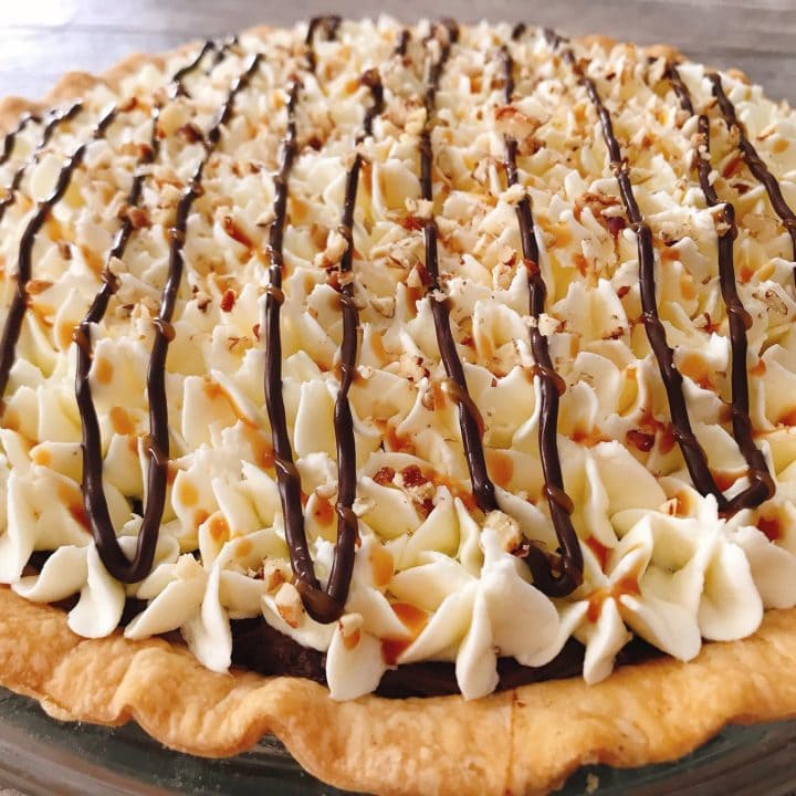 A close up of a Caramel Pecan Silk Pie with whipped cream drizzled with melted chocolate, caramel sauce, and sprinkled with chopped pecans.