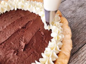 Whipped cream being piped over the chocolate layer of a Caramel Pecan Silk Pie.