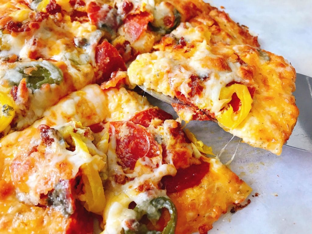 A delicious Keto Pizza topped with peppers and pepperoni with a slice being lifted with a spatula.