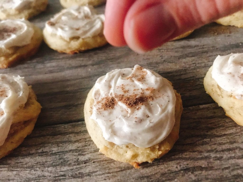 Fingers sprinkling ground nutmeg over frosted eggnog Christmas cookies.