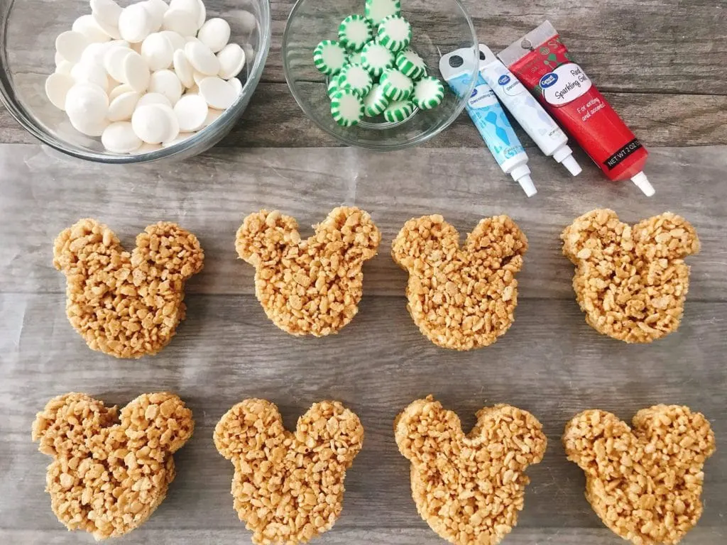 A bowl of marshmallows, a bowl of green starlight mints, decorating gel, and 8 Mickey Mouse shaped Gingerbread Rice Krispie Treats.