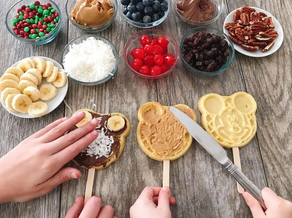 Waffle bar toppings in bowls, two kids adding toppings to Mickey Waffles to make a Mickey Mouse Waffle Bar