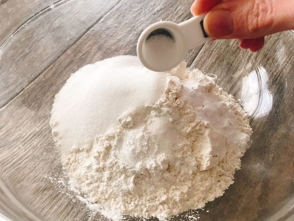 A measuring spoon pouring salt into a bowl of dry ingredients to make Sweet Cream Waffles.