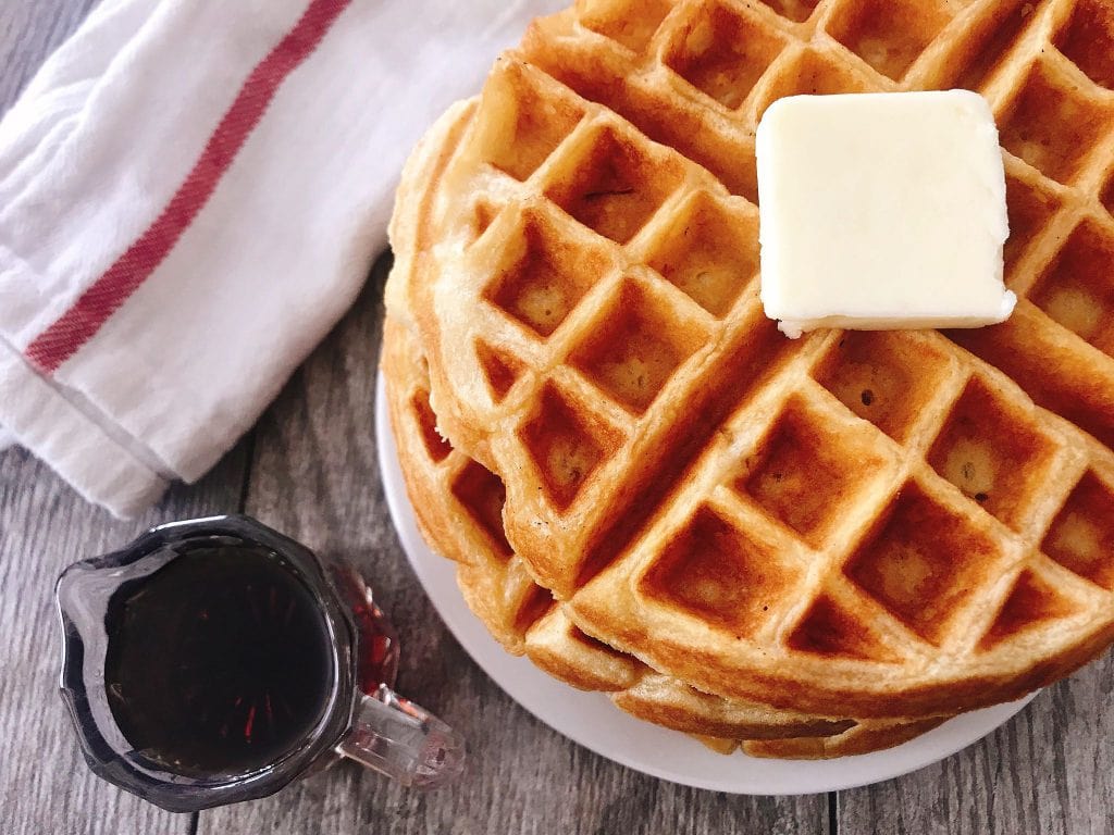 A stack of waffles with a pat of butter and a cup of syrup.