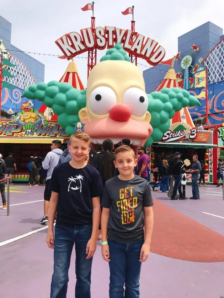 Two kids standing in front of a large clown with a sign that says “Krustyland”