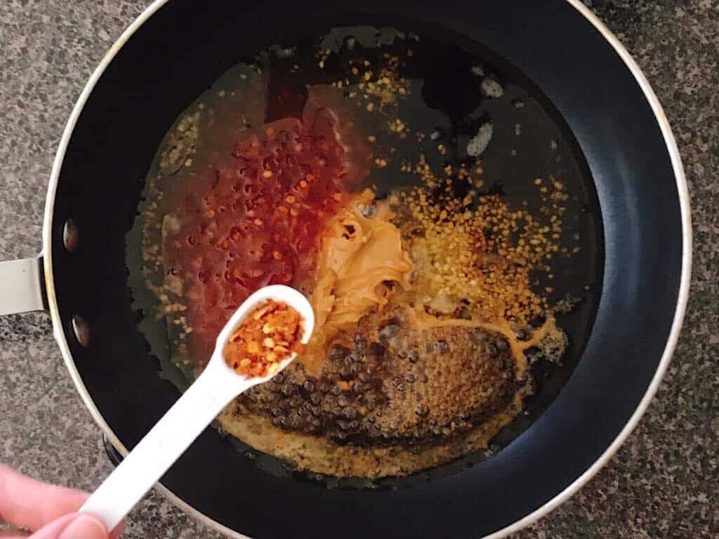 Red pepper flakes in a measuring spoon about to be added to a saucepan.