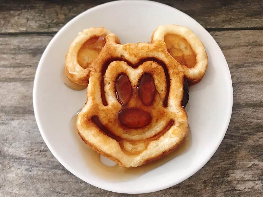 A Mickey Mouse shaped crispy waffle with syrup on a white plate.