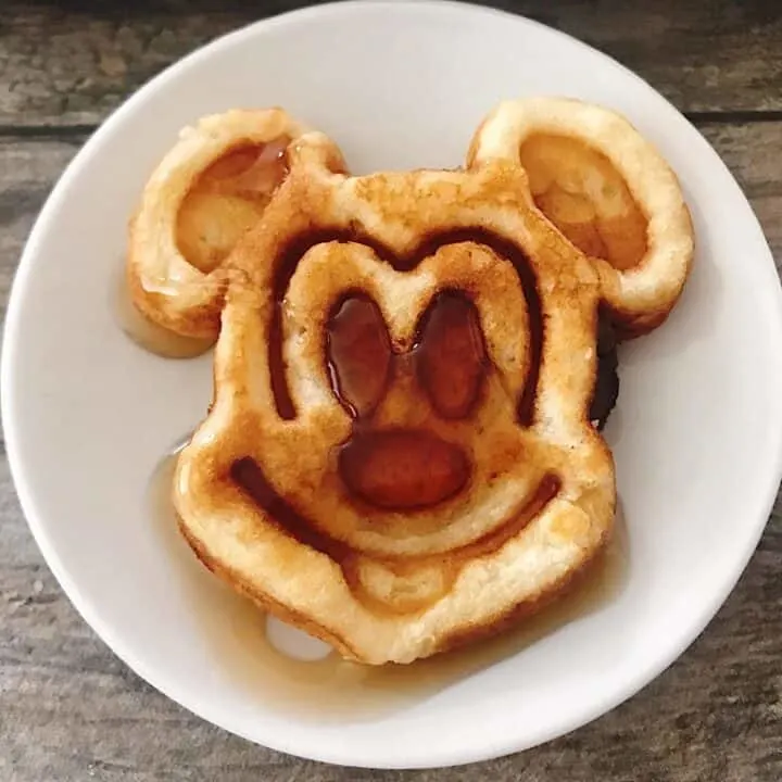 A Mickey Mouse shaped crispy waffle with syrup on a white plate.