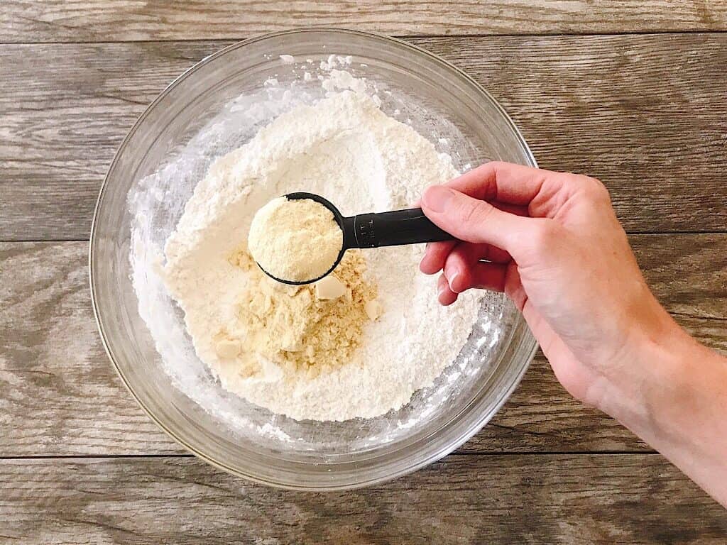 A measuring spoon of malted milk over a bowl of crispy waffle batter dry ingredients.