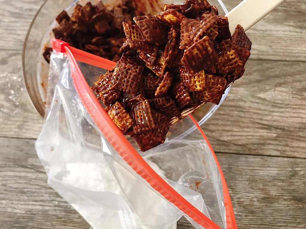 A spoon scooping A bag of chocolate covered Chex cereal in a ziplock bag with powdered sugar.