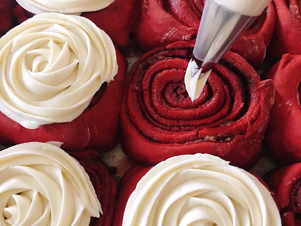 Cream cheese frosting being piped onto red velvet cinnamon rolls.