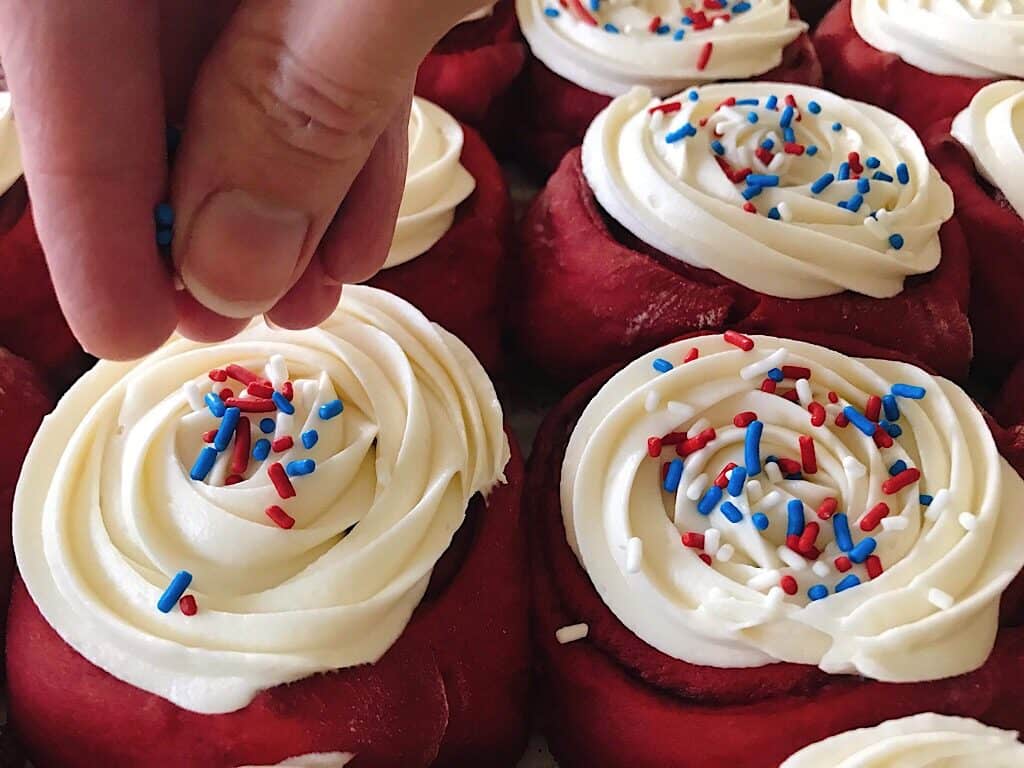 Sprinkles on red velvet cake mix cinnamon rolls with cream cheese frosting.