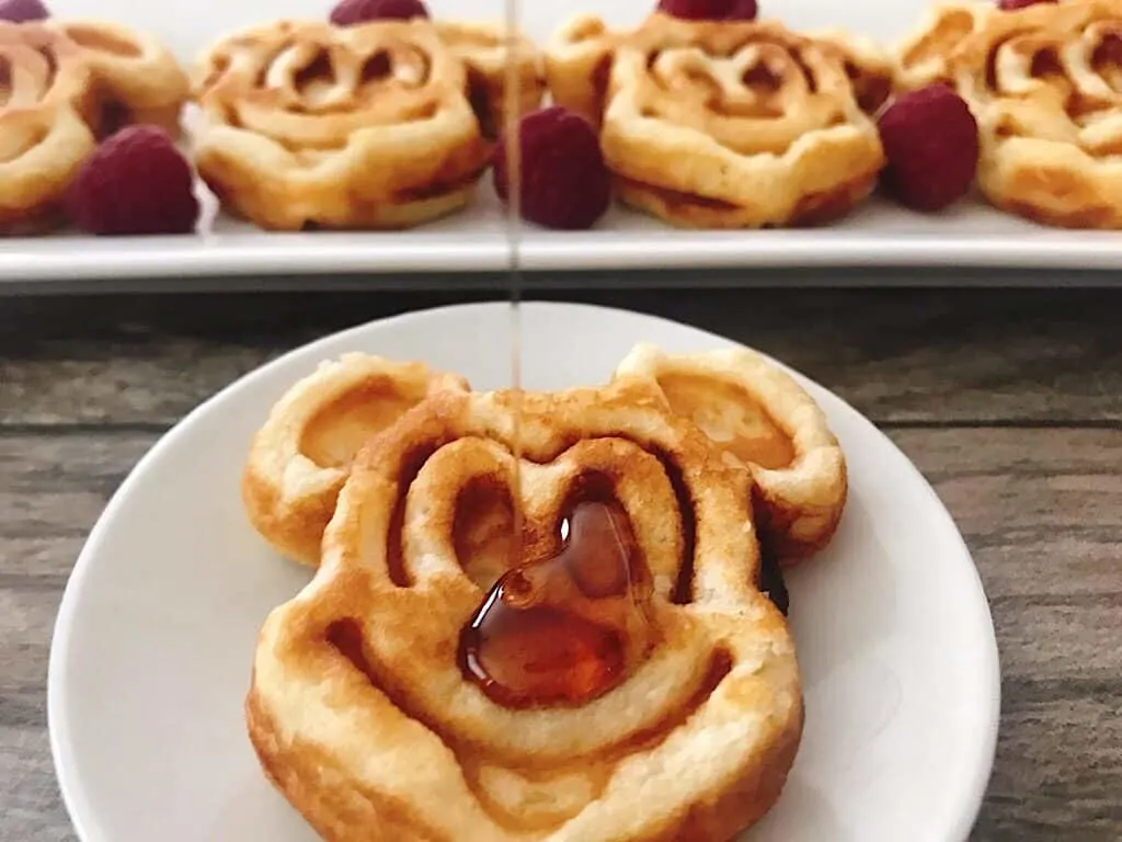 Crispy Mickey Mouse shaped waffles lined up on a white plate with raspberries.