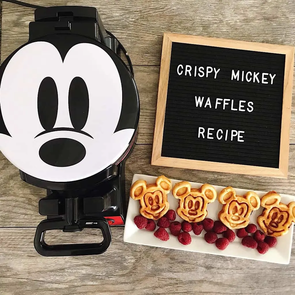 A waffle iron with a Mickey Mouse face and a letter board that says “crispy Mickey Waffles Recipe” and a plate of Mickey Mouse shaped waffles on a white plate with raspberries.