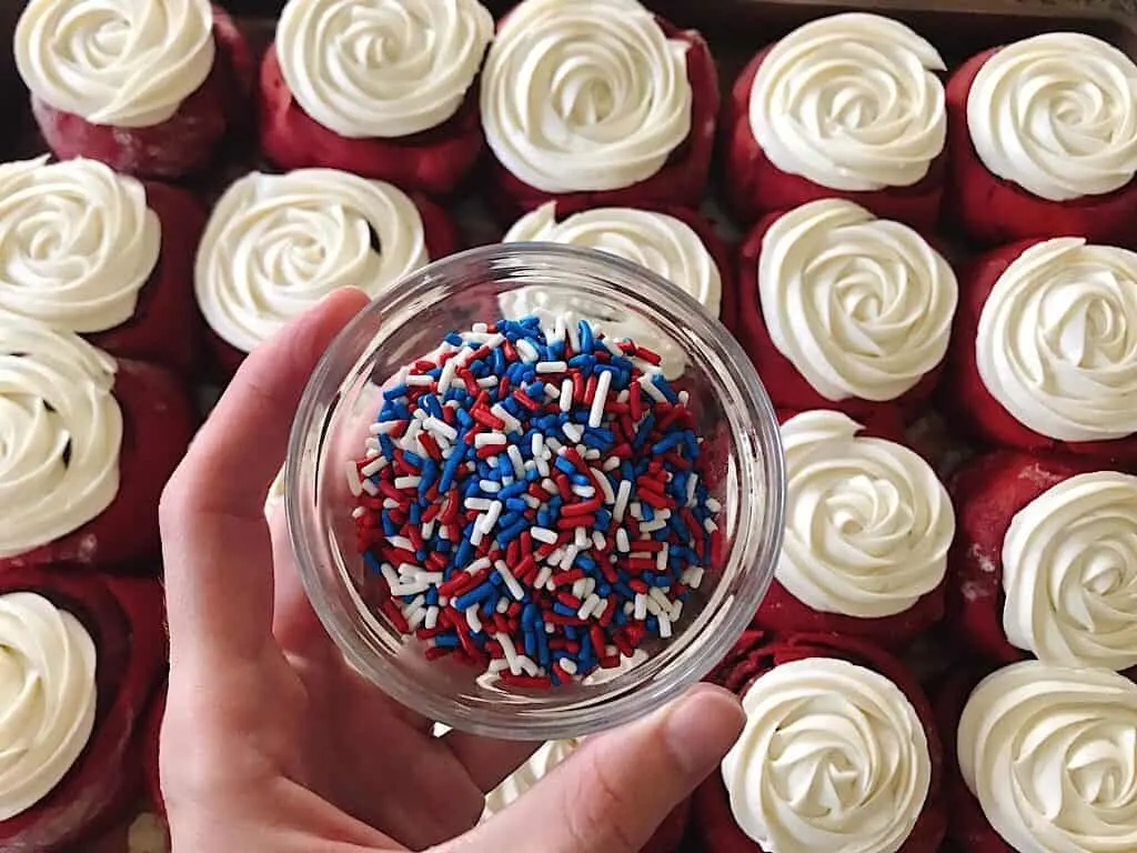 A bowl of red, white, and blue sprinkles over a tray of red velvet cinnamon rolls.