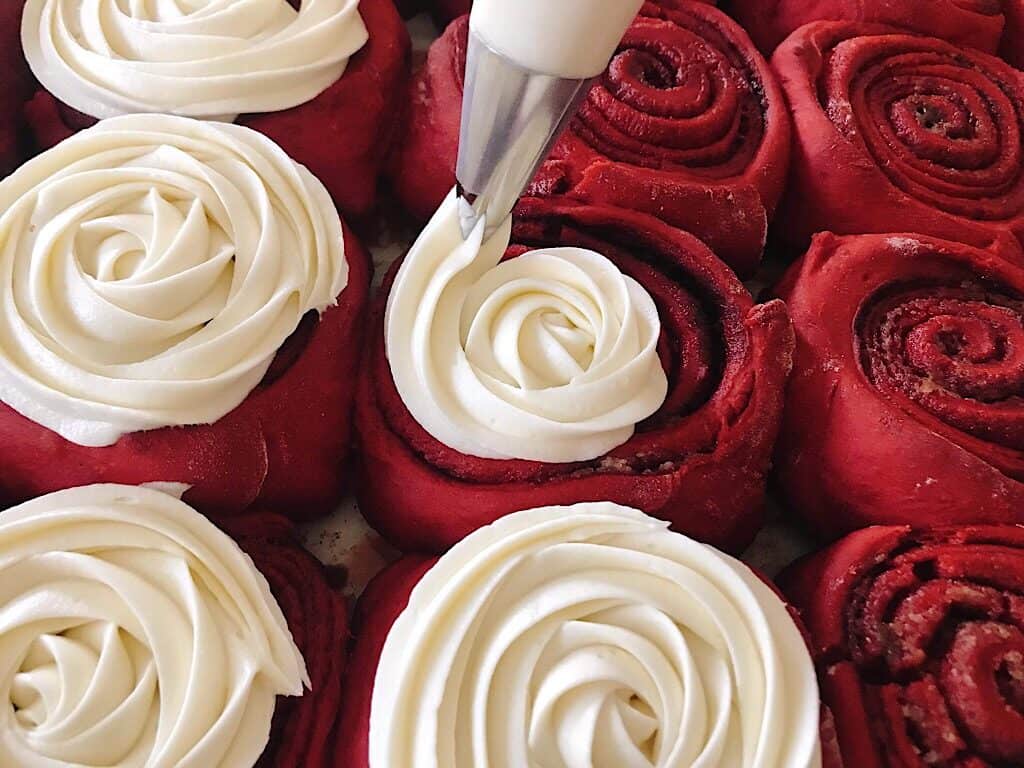 Cream cheese frosting being piped onto red velvet cinnamon rolls.