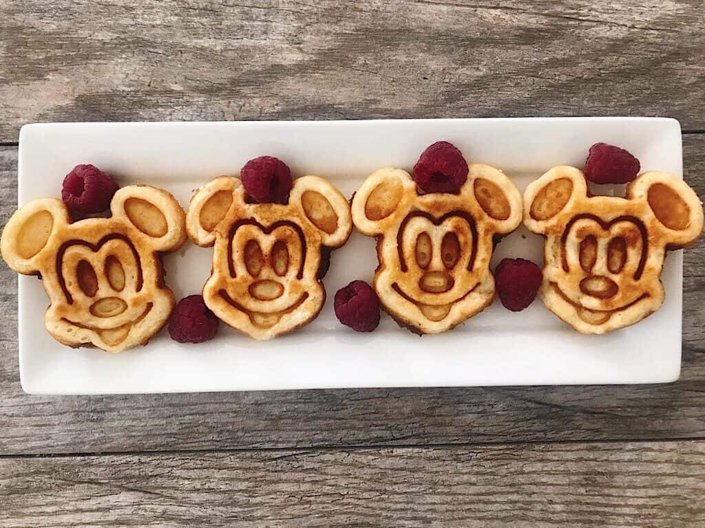 Crispy Mickey Mouse shaped waffles lined up on a white plate with raspberries.