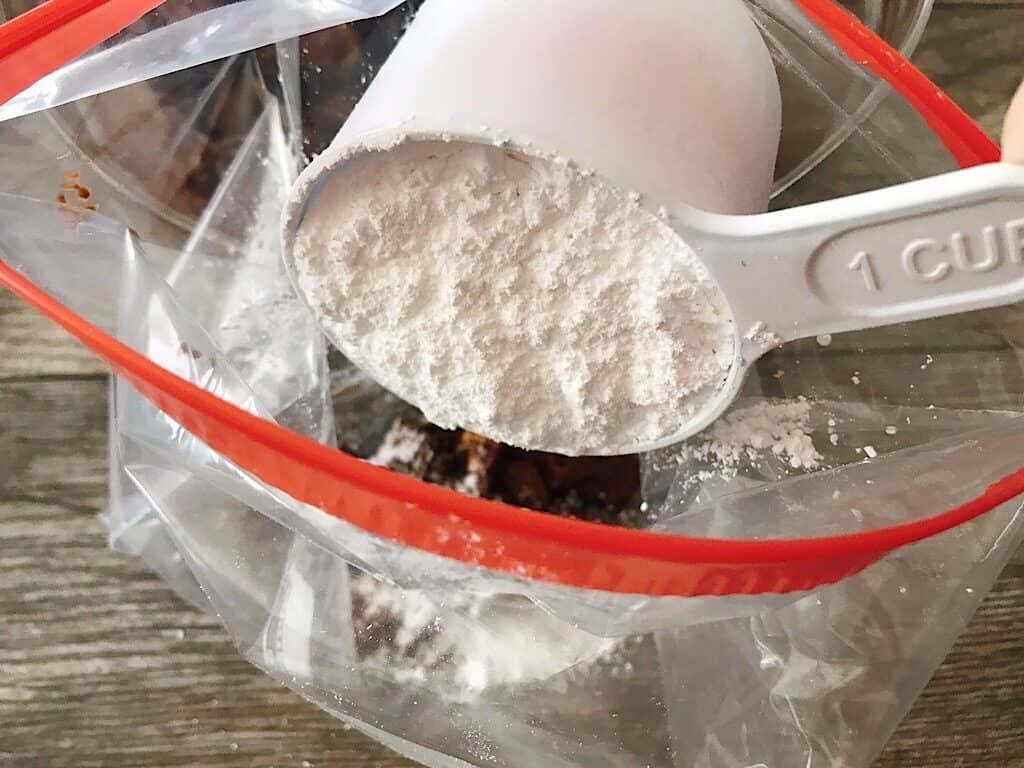 A measuring cup pouring powdered sugar into a ziplock bag.