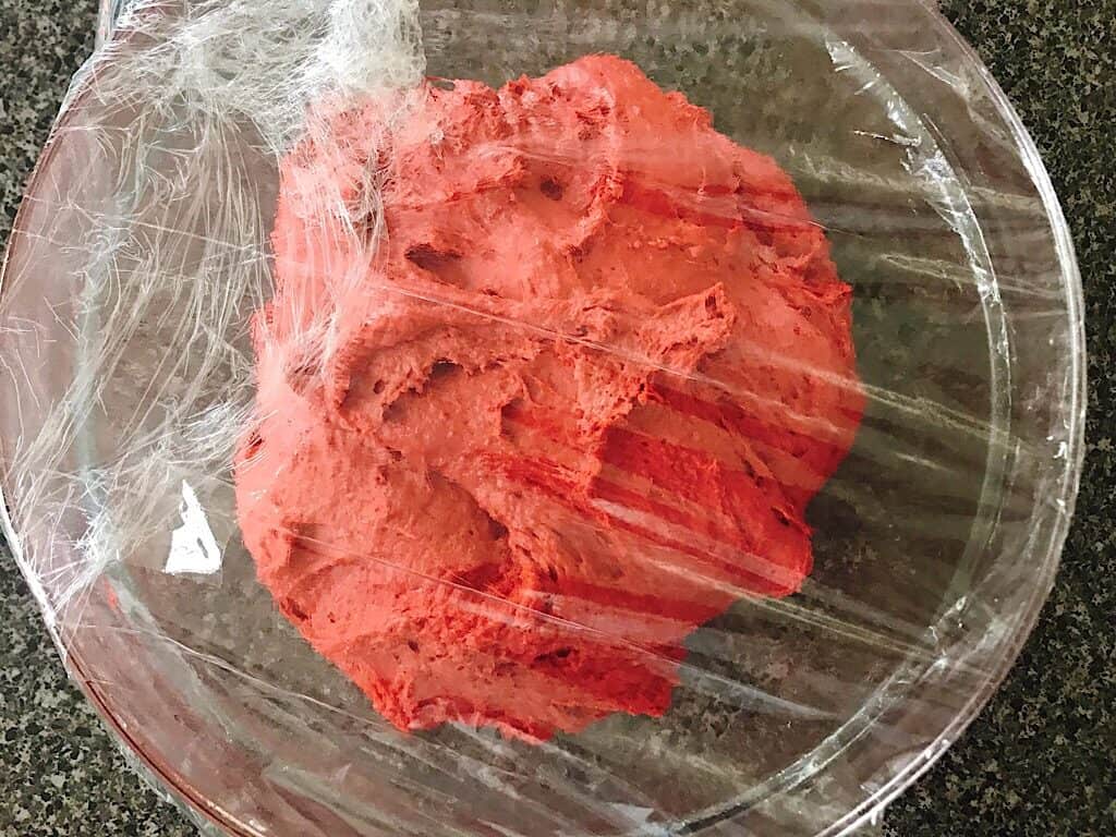 Red velvet Cinnamon Roll dough in a bowl covered with plastic wrap.