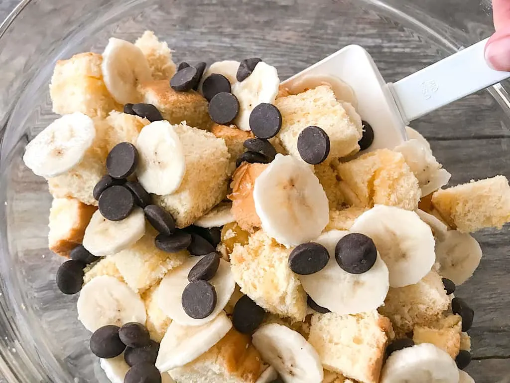 Bread cubes, bananas, and chocolate chips being mixed in a bowl to make Chocolate Peanut Butter Banana French Toast Bake