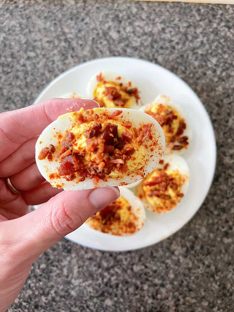 A had holding a deviled egg topped with bacon over a plate of crack deviled eggs.
