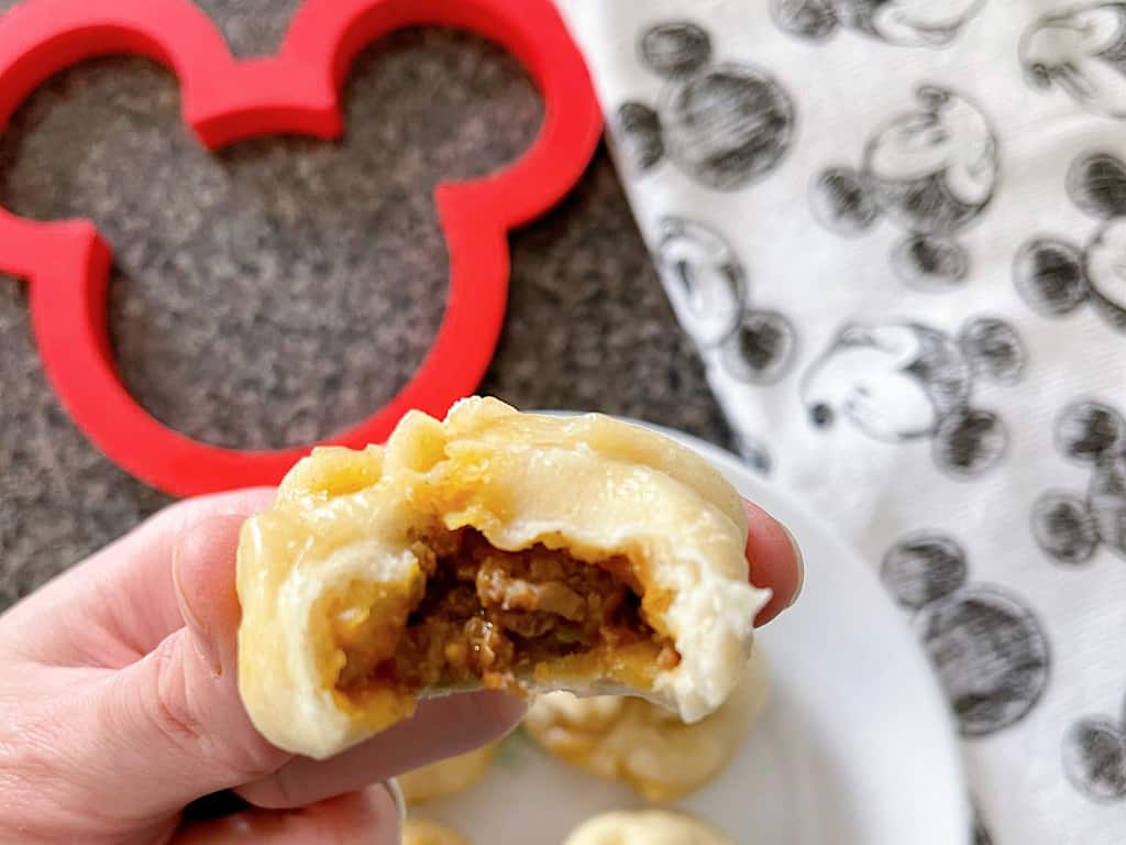 A hand holding a cheeseburger bao bun in front of a red Mickey Mouse silhouette