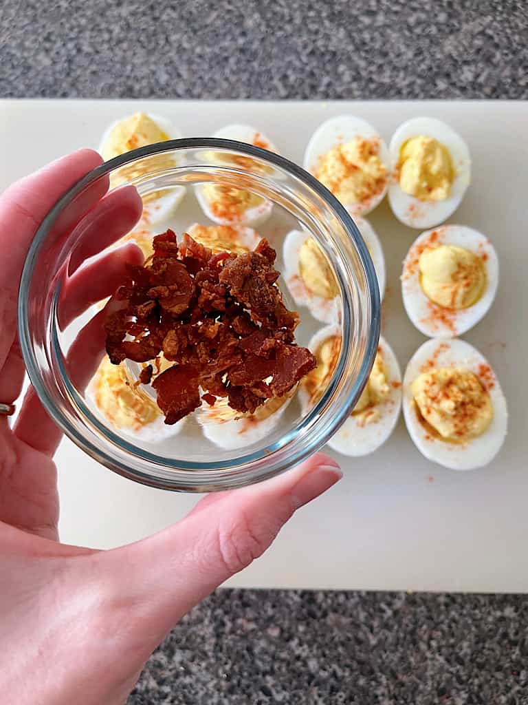 Bacon pieces in a bowl held over crack deviled eggs.