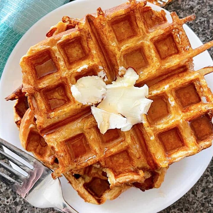 French Toast Waffles on a plate topped with butter next to a fork and a blue and white towel.