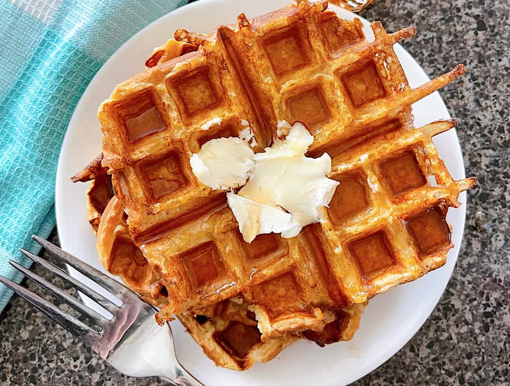 French Toast Waffles on a plate topped with butter next to a fork and a blue and white towel.