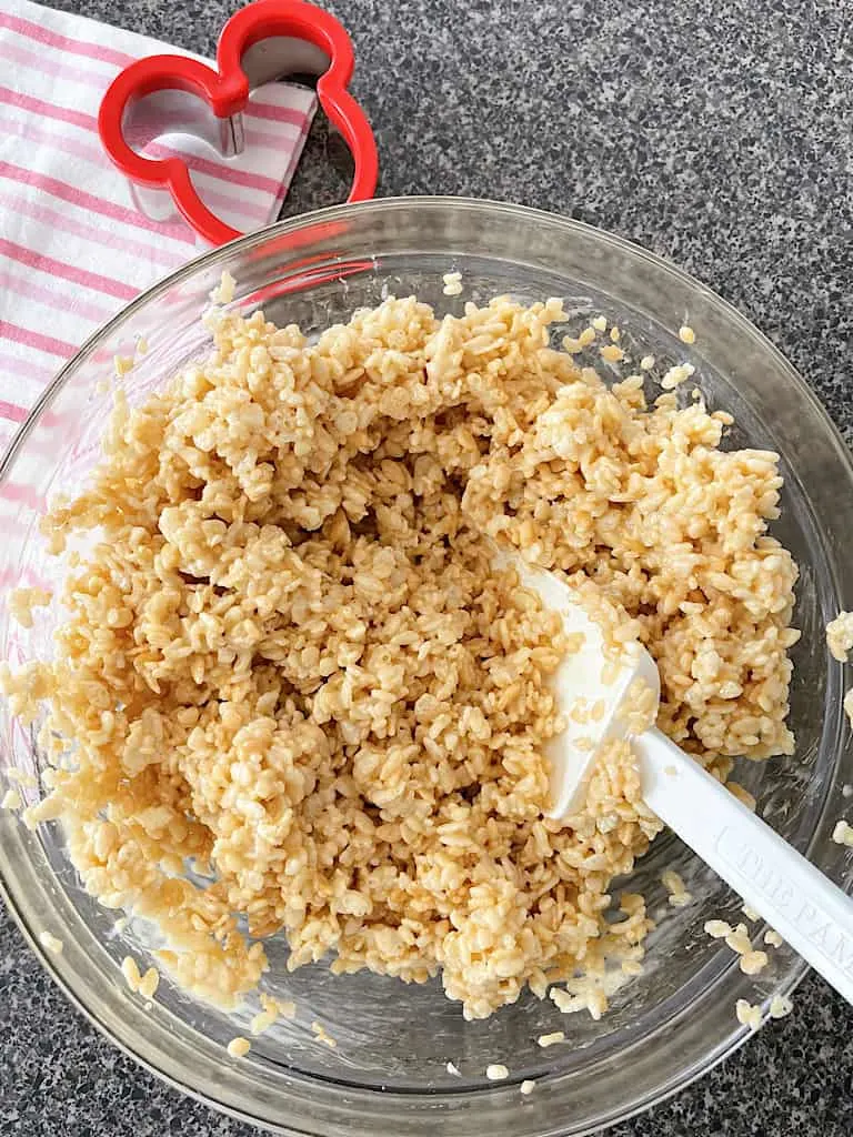 A bowl of Rice Krispies mixed with melted marshmallows next to a pink and white dish towel and a Mickey Mouse cookie cutter.