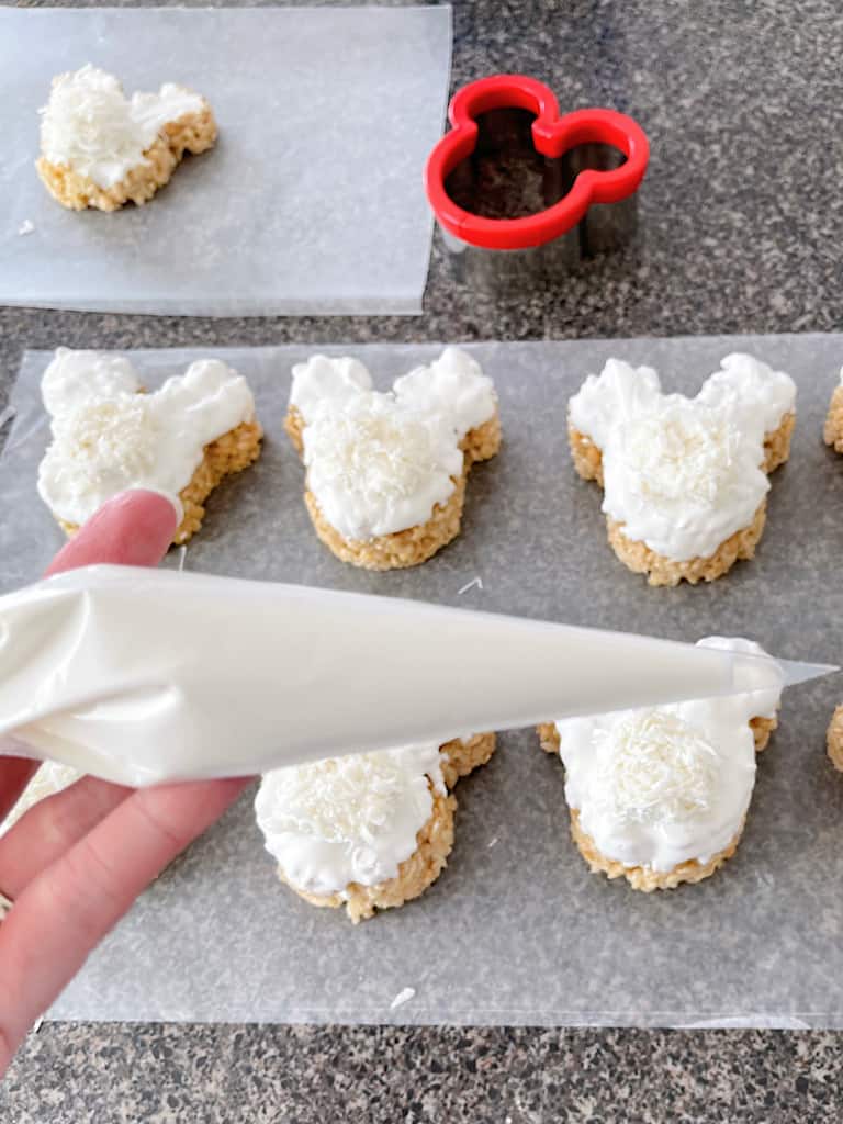 A piping bag full of melted white candy melts over Mickey Rice Krispie Bunny treats.
